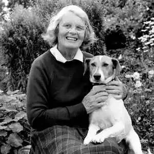 Black and white photo of a smiling elderly woman with glasses holding a white and brown dog in a garden setting