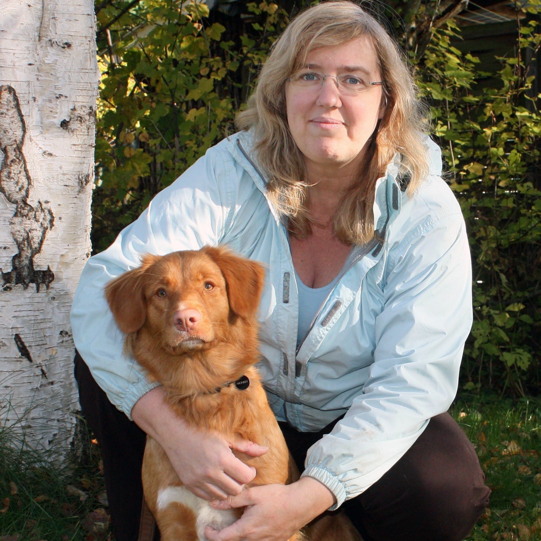 Woman in light jacket kneeling outdoors with a brown dog beside a birch tree and green foliage