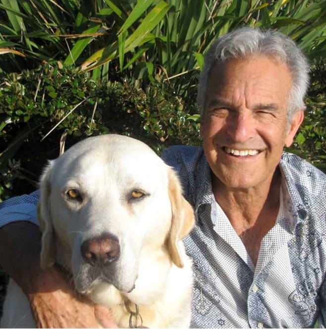Smiling elderly man with gray hair sitting outdoors with large white Labrador dog near green plants