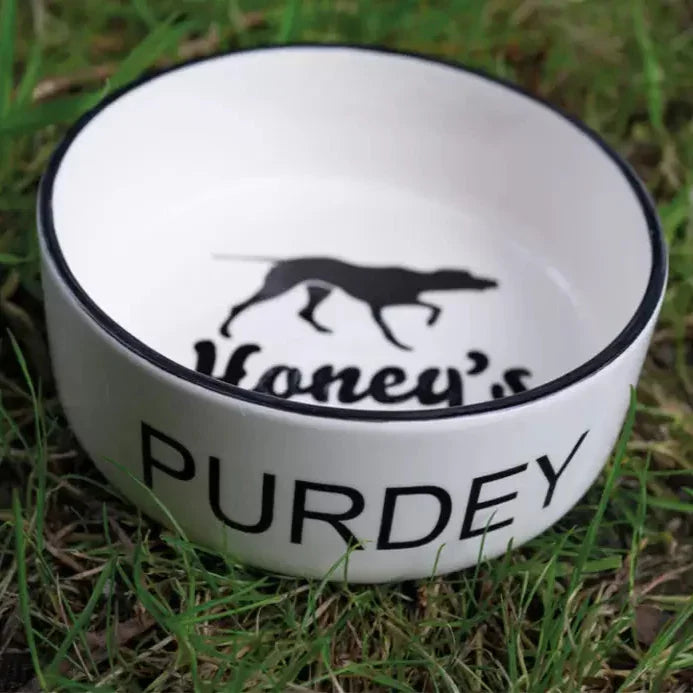 White ceramic dog bowl labeled "Purdey" with black greyhound silhouette and "Honey's" text inside, on grass