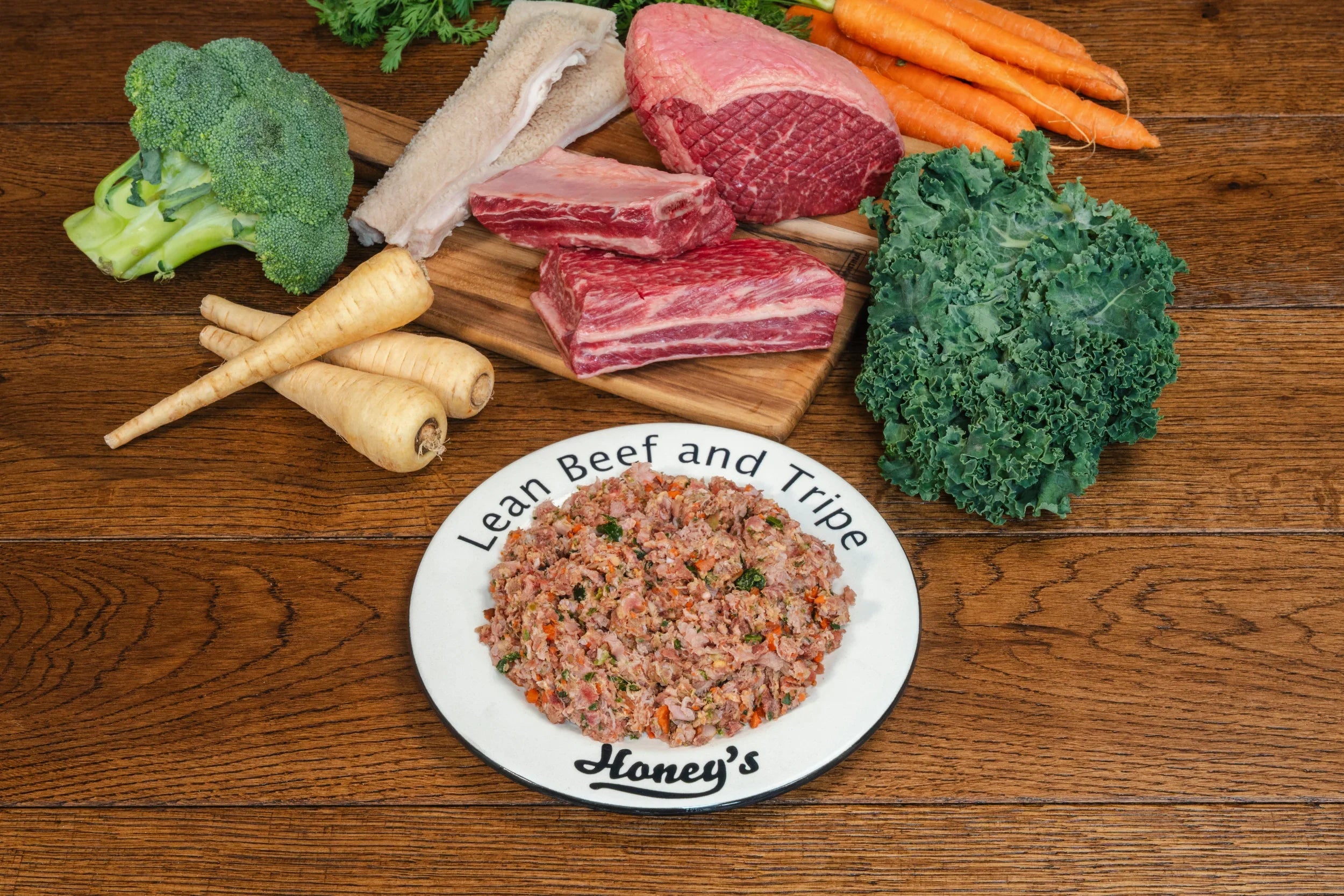 Lean beef and tripe dog food on plate with raw beef, tripe, broccoli, kale, carrots, and parsnips on wooden table